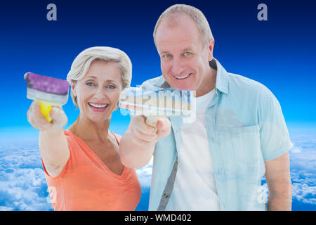 Happy older couple holding paintbrushes against mountain peak through the clouds Stock Photo