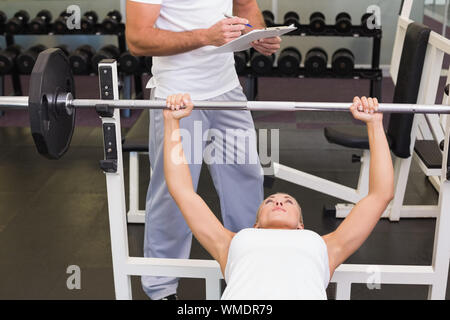Male trainer with clipboard besides young woman lifting barbell in the gym Stock Photo