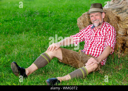 handsome bavarian man sitting outdoors at tree stump and smiles Stock Photo