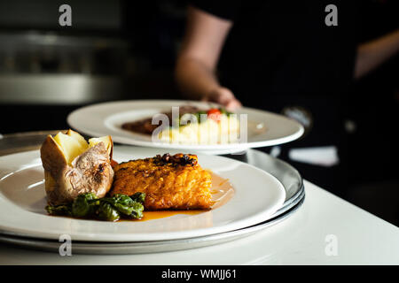 a plate with battered cod garnished with a roasted potato, some fried cherry tomatoes and some fried green peppers, in a restaurant, and a woman with Stock Photo