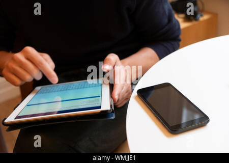 closeup of a young caucasian indoors using a digital tablet sitting at a white table, with a smartphone in the foreground Stock Photo