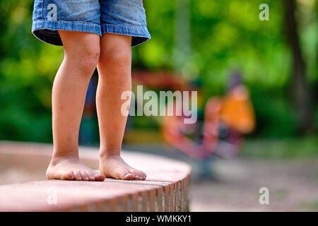 Low section of a caucasian child girl in jeans shorts who is standing barefoot on a little stone wall with sandy feet in summer Stock Photo