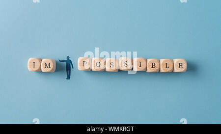 Wide view image of a silhouetted man figure pushing away letters IM from the word Impossible spelled on wooden dices. Over blue background. Stock Photo