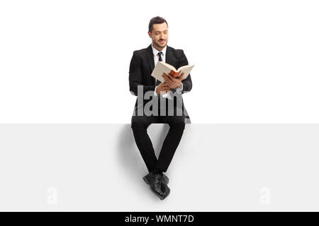 Full length portrait of a man in a suit sitting on a blank banner and reading a book isolated on white background Stock Photo