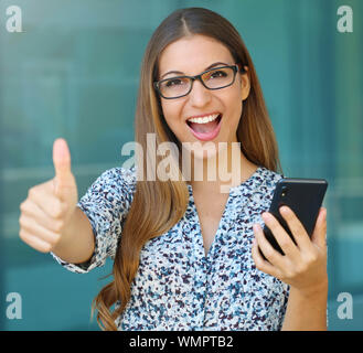 Successful joyful business woman with thumb up holds smart phone and looks at camera. Stock Photo