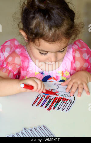 Curly brunette cute little toddler girl painting with color pen paper menorah and candle Jewish holiday Chanukah. Hebrew text : Happy Hanukkah. Stock Photo