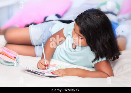 A 8 year old girl wearing denim shorts doing her math homework while laying in her comfy bed with stuffed animals in background. Stock Photo
