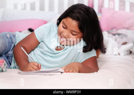 A young elementary school student with a positive attitude, doing her homework with pencil in hand; while laying in bed. Stock Photo