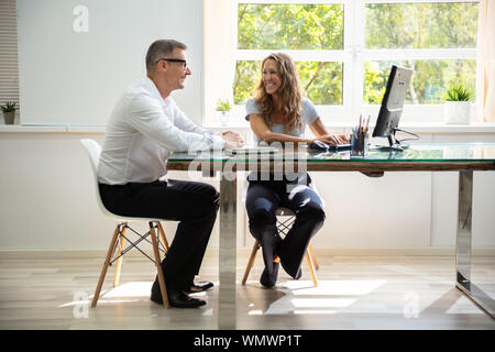 Smiling Male And Female Colleagues Sitting At Office Desk Working Together While Using Computer Stock Photo