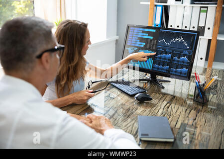 Side View Of Two Stock Market Brokers Discussing Graphs On Computer At Workplace Stock Photo