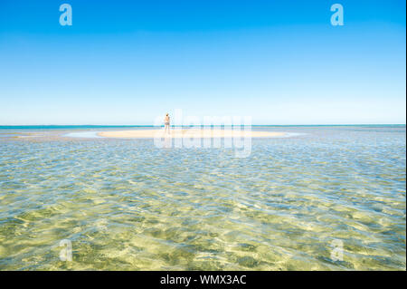 Distant view of solitary figure in a straw hat standing on a sand bar above the calm shallow waters of a sunny tropical island beach Stock Photo