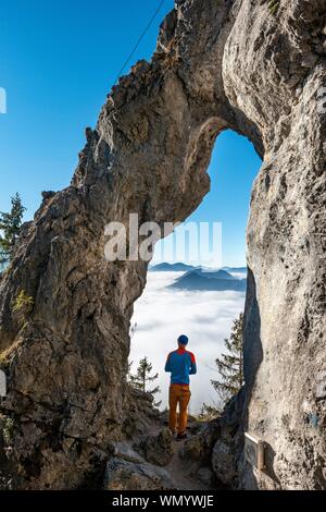 Young man stands in the rock gate Breitensteinfensterl with a view of the high fog above the valley, hiking trail to Breitenstein, Fischbachau Stock Photo