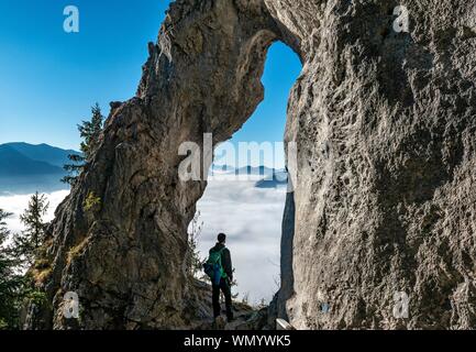 Young man stands in the rock gate Breitensteinfensterl with a view of the high fog above the valley, hiking trail to Breitenstein, Fischbachau Stock Photo