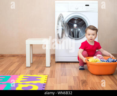 Little girl with a washbowl full of clothes in front of the washing machine Stock Photo