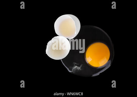Broken white raw egg, yellow round yolk, two cracked halves of eggshell on black background isolated close up top view, unusual design, Easter banner Stock Photo