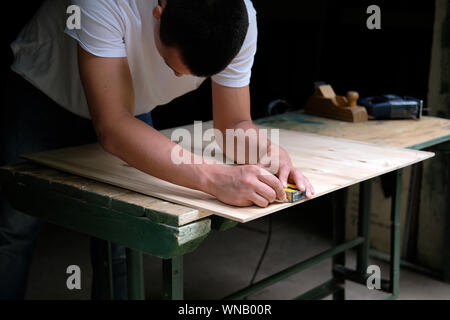 Carpenter marking straight line on plywood sheet using spirit level in carpentry workshop. Measuring, drawing line, DIY concept. Stock Photo