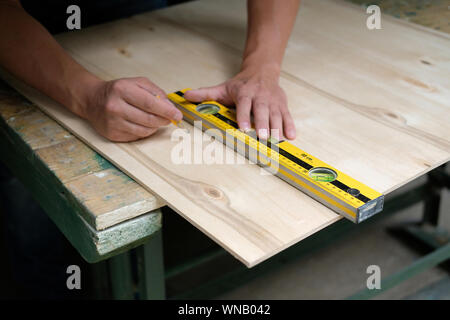 Carpenter marking straight line on plywood sheet using spirit level in carpentry workshop. Measuring, drawing line, DIY concept. Stock Photo