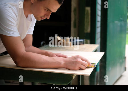 Carpenter marking straight line on plywood sheet using spirit level in carpentry workshop. Measuring, drawing line, DIY concept. Stock Photo