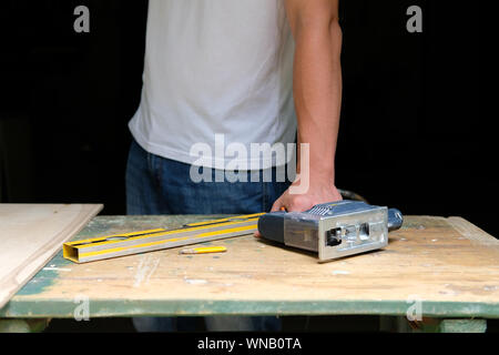 Hands of human holding electric jigsaw lying on workbench with spirit level and yellow pen. DIY concept. Stock Photo