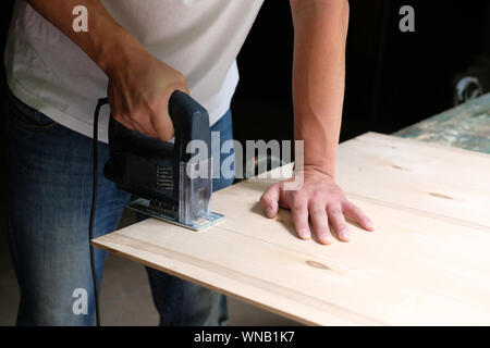 Carpenter is sawing a plywood sheet with electric jig saw machine in carpentry workshop. Close up hand with jigsaw. Carpentry concept. Stock Photo