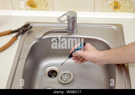 Close-up of handsome plumber repairing drain of kitchen sink. Photo of metalic sink and handyman with screwdriver Stock Photo
