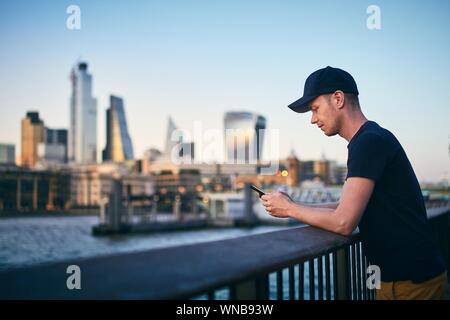 Side view of young man while using smart phone against London skyline at dusk. Stock Photo