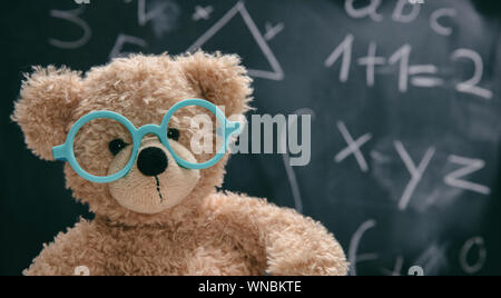 Maths concept. Smart kid in school class, cute teddy wearing blue eyeglasses and math symbols on blackboard Stock Photo