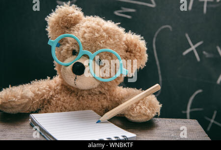 Maths lesson test concept. Smart kid in school class, cute teddy wearing blue eyeglasses and math symbols on blackboard Stock Photo
