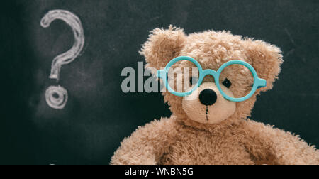 Confused child, doubt. Smart kid in class, question mark on blackboard, cute teddy wearing blue eyeglasses Stock Photo