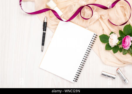 Flat lay paper notebook, roses flowers, ribbon, beige scarf, lipstick on wooden desk. Top view woman home office workspace, feminine background. Fashi Stock Photo