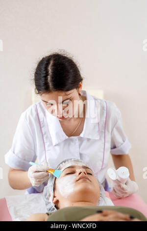 Cosmetology specialist applying facial mask using brush, making skin hydrated and healthy. Attractive woman relaxing. Beautician at work. Hands of cosmetology. Stock Photo