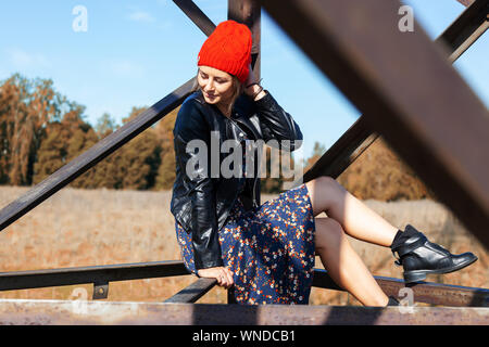 Portrait of a  cheerful woman in a knitted hat,  leather coat, pretty dress and boots, smiles cute, poses, smiles and sits on birch logs. The concept Stock Photo