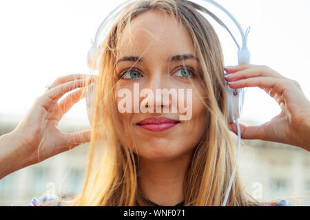 Beautiful young blonde woman with long hair and blue eyes listening to music with white headpones outside. Hands on headphones. Rome, Italy, Sunset. Stock Photo