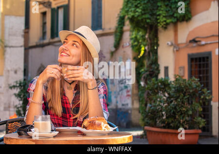 Young blonde woman with blue eyes having breakfast in typical Italian bar outside in historical neighborhood Trastevere in Rome, Italy. Cappuccino Stock Photo