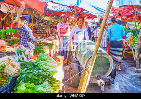 TAUNGGYI, MYANMAR - FEBRUARY 20, 2018: The porter moves the wheelbarrow with large barrel through the narrow alley of agricultural market, lined with Stock Photo