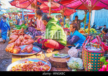 TAUNGGYI, MYANMAR - FEBRUARY 20, 2018: The butchery stalls of agricultural market with vendors, sitting on the tables next to the trays with chicken, Stock Photo