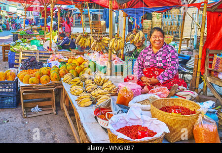 TAUNGGYI, MYANMAR - FEBRUARY 20, 2018: The smiling fruit vendor, sitting at the bunches of bananas and bbaskets with chili pepper in a stall of agricu Stock Photo