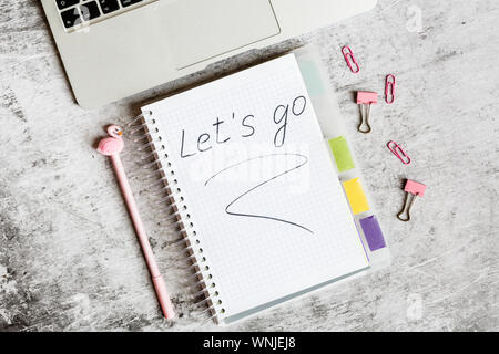 Desktop flatlay items: laptop, notebook with inscription Let is go, pink pen with flamingo, paper clips lying on gray marble background Stock Photo