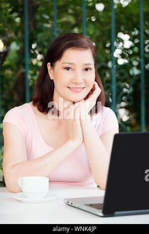 Portrait of positive mature female freelancer sitting at cafe table with cup of coffee and working on laptop Stock Photo