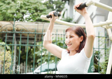 Happy middle-aged Asian woman doing exercises in lat pull down machine outdoors Stock Photo