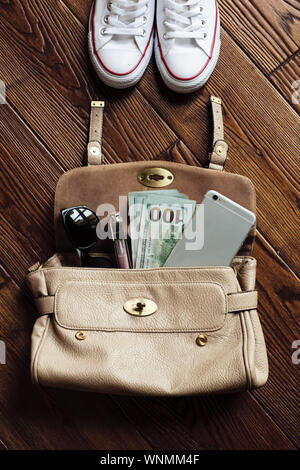 Top view contents of women bag. Layout lipgloss, cash money, mobile phone and sunglasses in a leather bag on brown wooden background Stock Photo