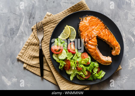 Fried salmon fish steak with vegetable salad and lemon on black plate. Creative layout made of fish, lettuce, tomato, cucumber, top view, flat lay Stock Photo