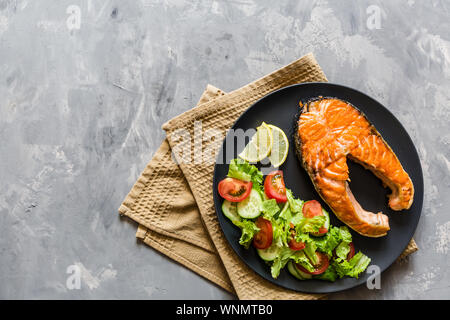 Fried salmon fish steak with vegetable salad and lemon on black plate. Creative layout made of fish, lettuce, tomato, cucumber, top view, flat lay Stock Photo