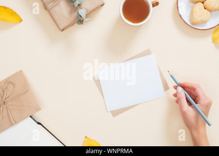 Woman hand writing letter over autumn background with fallen leaves, cup of tea, cookies, gift box. Autumn composition, flat lay, top view, overhead Stock Photo