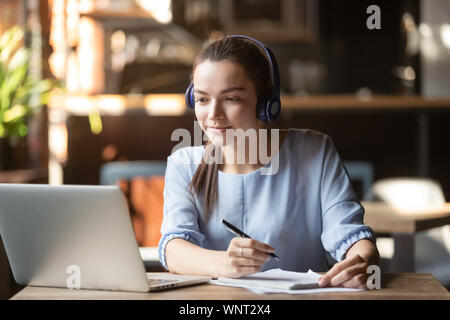 Focused woman wearing headphones using laptop, writing notes Stock Photo