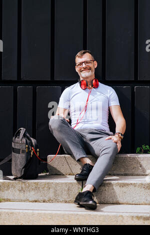 Bearded mature man sitting on steps with headphones while relaxing on a sunny day Stock Photo
