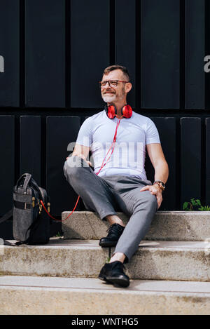 Bearded mature man sitting on steps with headphones while relaxing on a sunny day Stock Photo