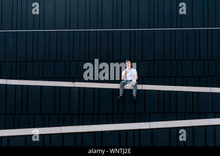 mature bearded businessman with smartphone sitting on wall while looking camera Stock Photo