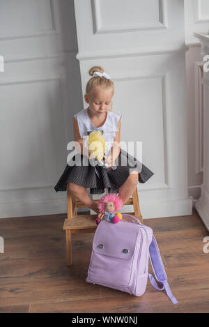 Close-up portrait of a little girl against a white wall.Girl in a school uniform with The girl holds dolls in her hands. A school backpack is on the f Stock Photo