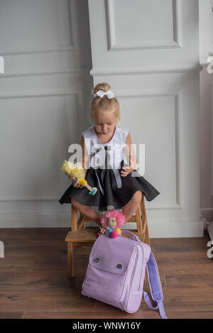 Close-up portrait of a little girl against a white wall.Girl in a school uniform with The girl holds dolls in her hands. A school backpack is on the f Stock Photo